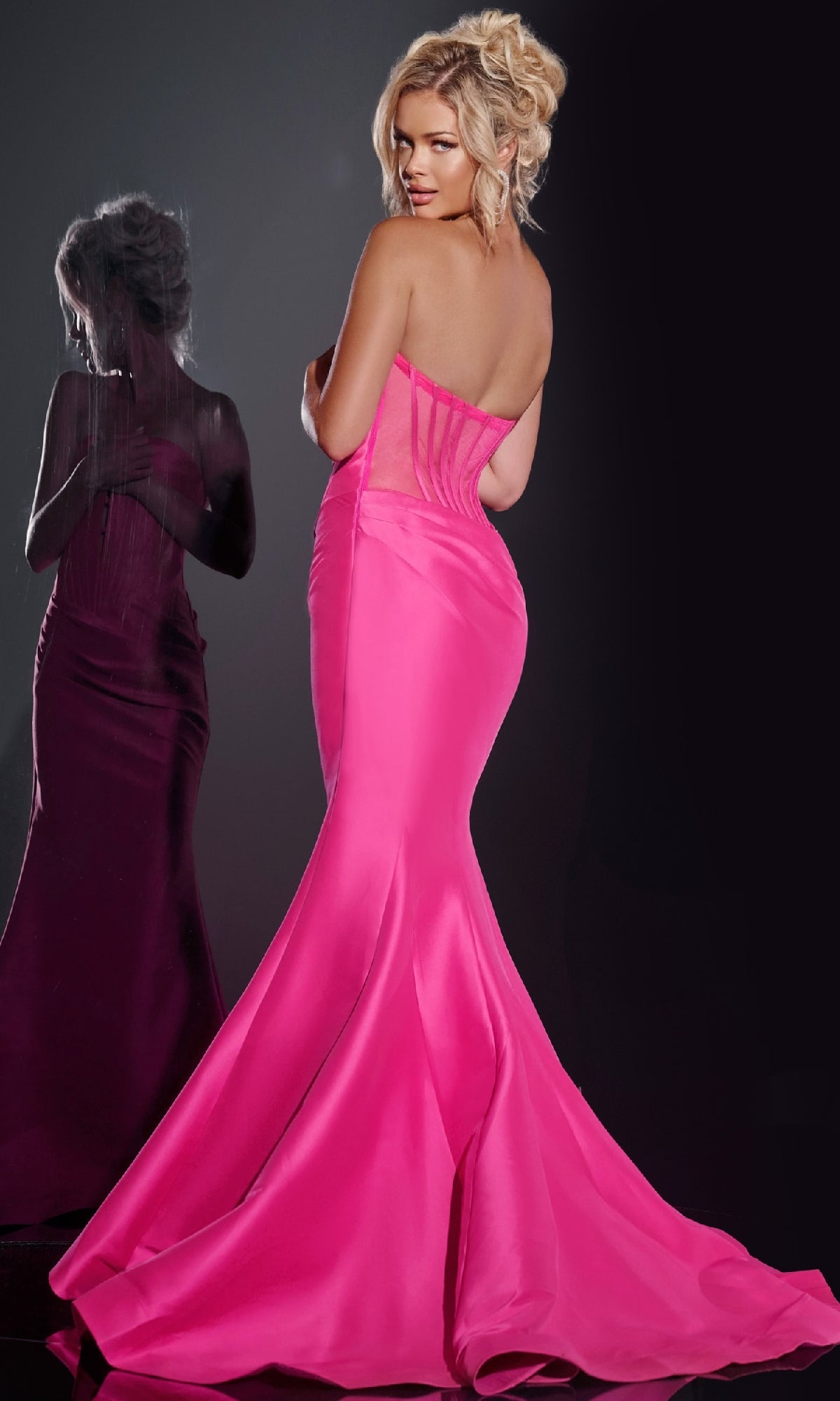 A woman in the Sheer-Back Strapless Prom Gown, Jovani 43350, poses with her back to the camera, facing a mirror reflecting her image.